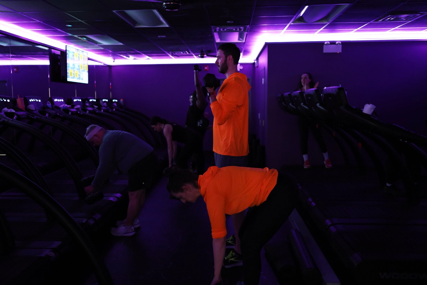 Group workout session at Momentum OP, featuring individuals in bright orange attire engaging in strength training with dumbbells in a modern gym environment, illuminated by purple lighting.
